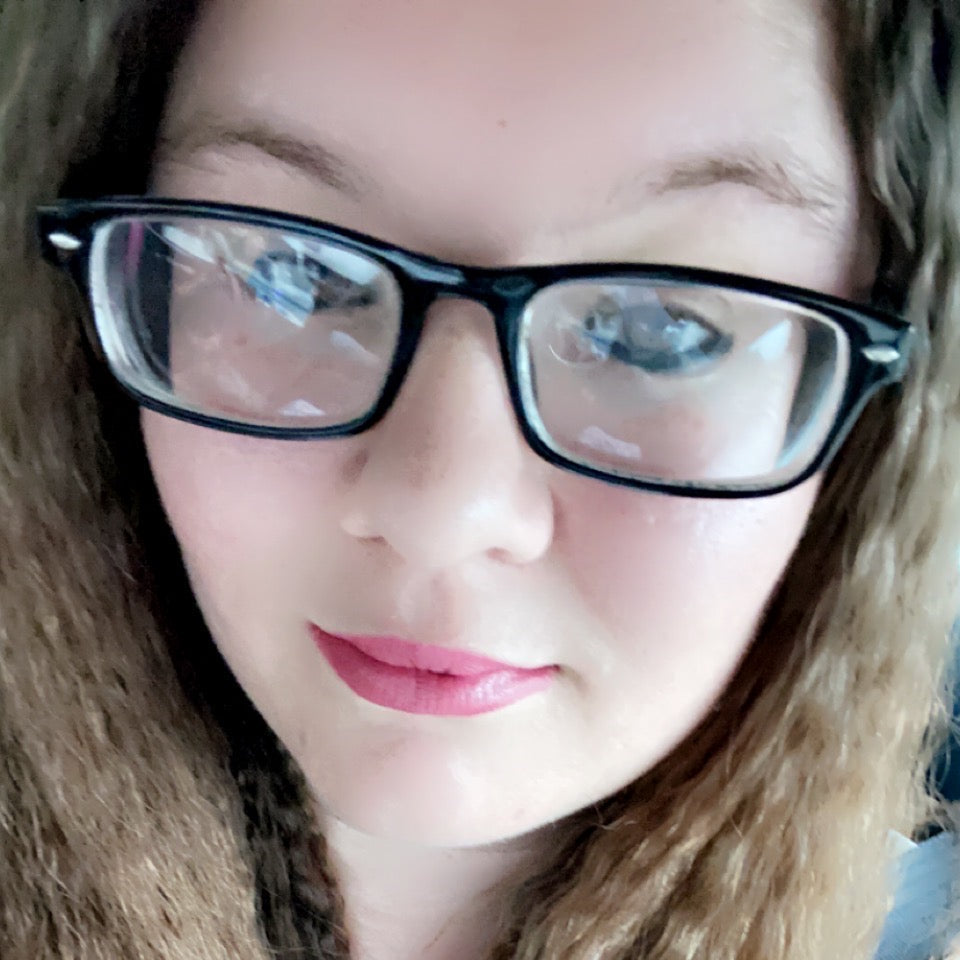 A close-up selfie of author Courtney Marie Valenzuela smiling, wearing thick black rectangular glasses, pink lipstick, and a white patterned shirt.