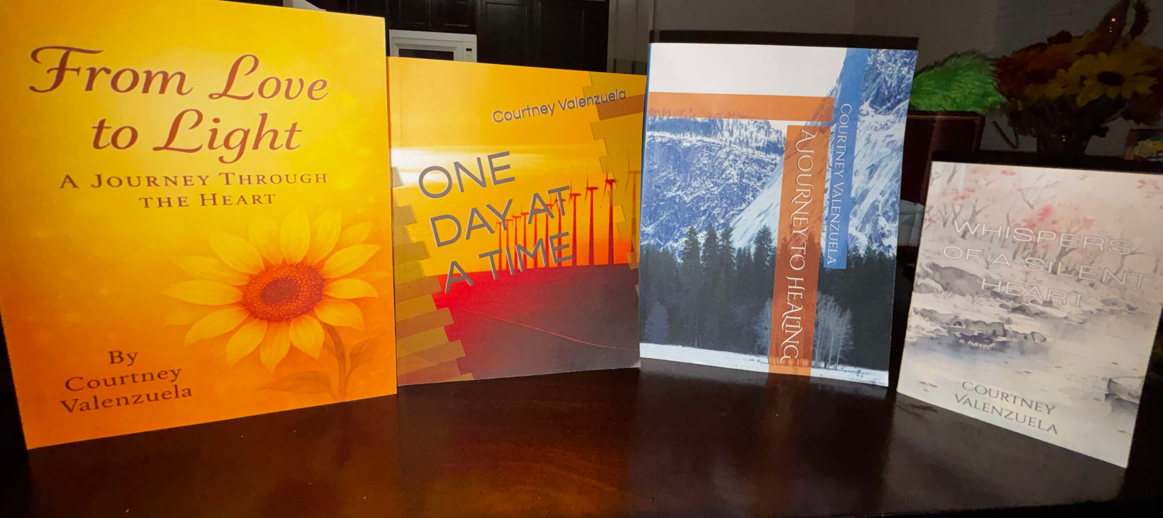 Four paperback books standing upright on a dark surface, featuring the titles "From Love to Light," "One Day at a Time," "A Journey to Healing," and "Whispers of the Heart" by author Courtney Marie Valenzuela.